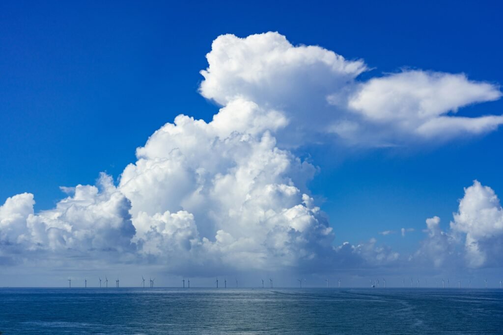 Scenic ocean landscape with towering clouds and distant wind turbines under a clear blue sky.